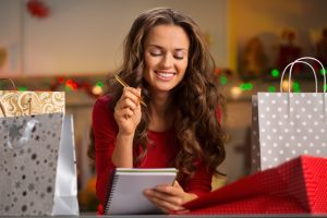Christmas holidays are a time of gift-giving. Shopping list can make Christmas season easy and save time for more fun. Happy young woman among shopping bagswith pen and notebook checking list